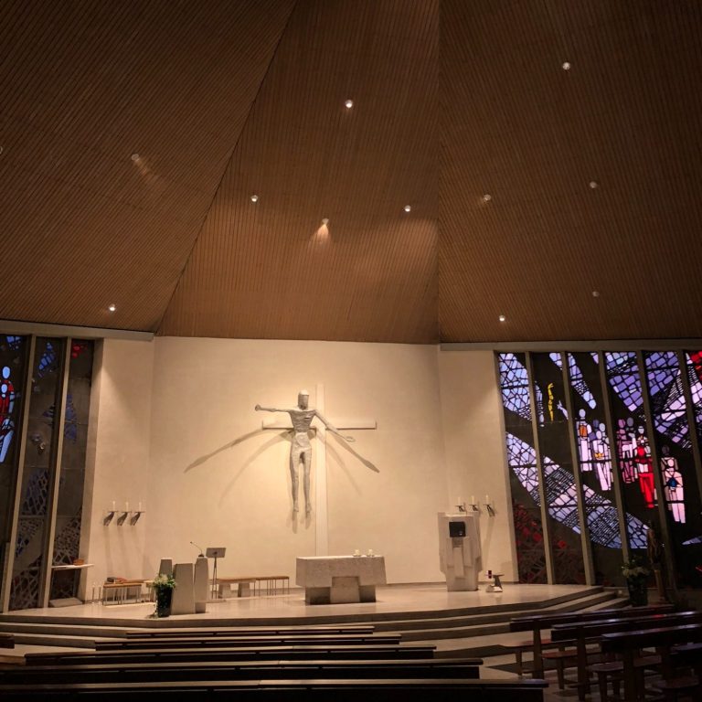 Moderne Kirche mit einem Christus-Kreuz und bunten Fenstern im Hintergrund.