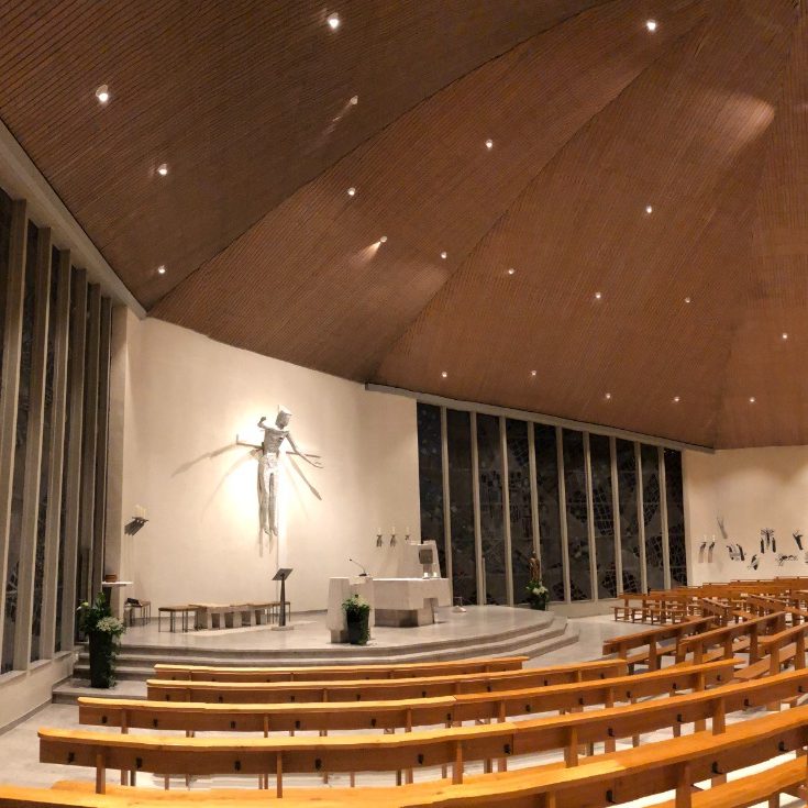Moderne Kirche mit hölzernem Gewölbe, Altar und Kruzifix. Bänke in halbkreisförmiger Anordnung.